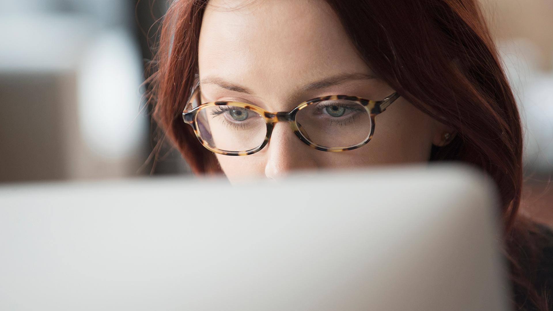 A woman in glasses, staring intently at a laptop screen.