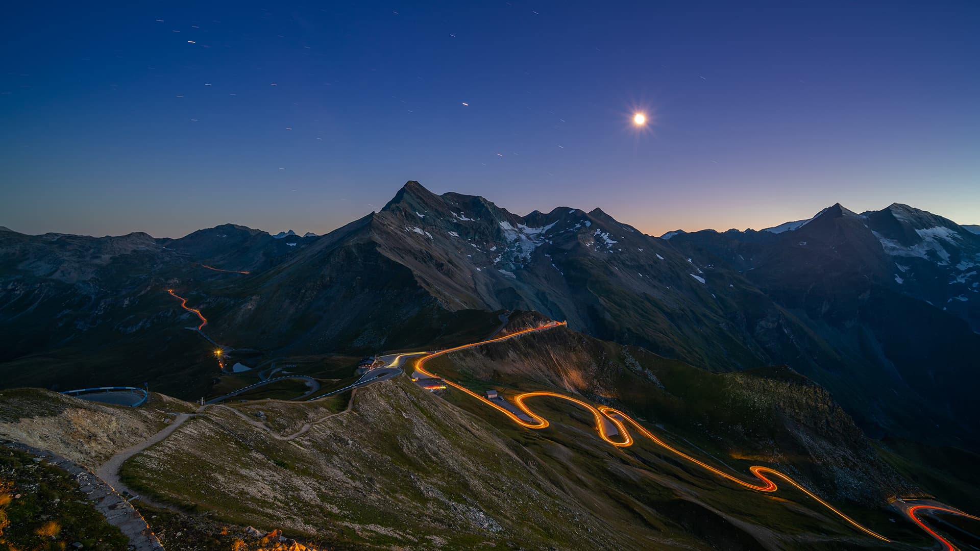 light trails on curvy high alpine road through european mountains at night with star shapped moon