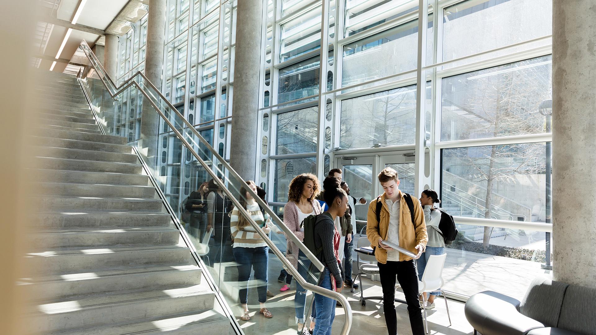 A group of male and female college students walk together to class. They are walking in a campus lobby or hallway lined with large, bright windows.