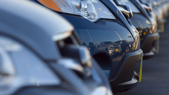 Row of brand new cars at a car dealership. Focus is on the headlight of a second car.