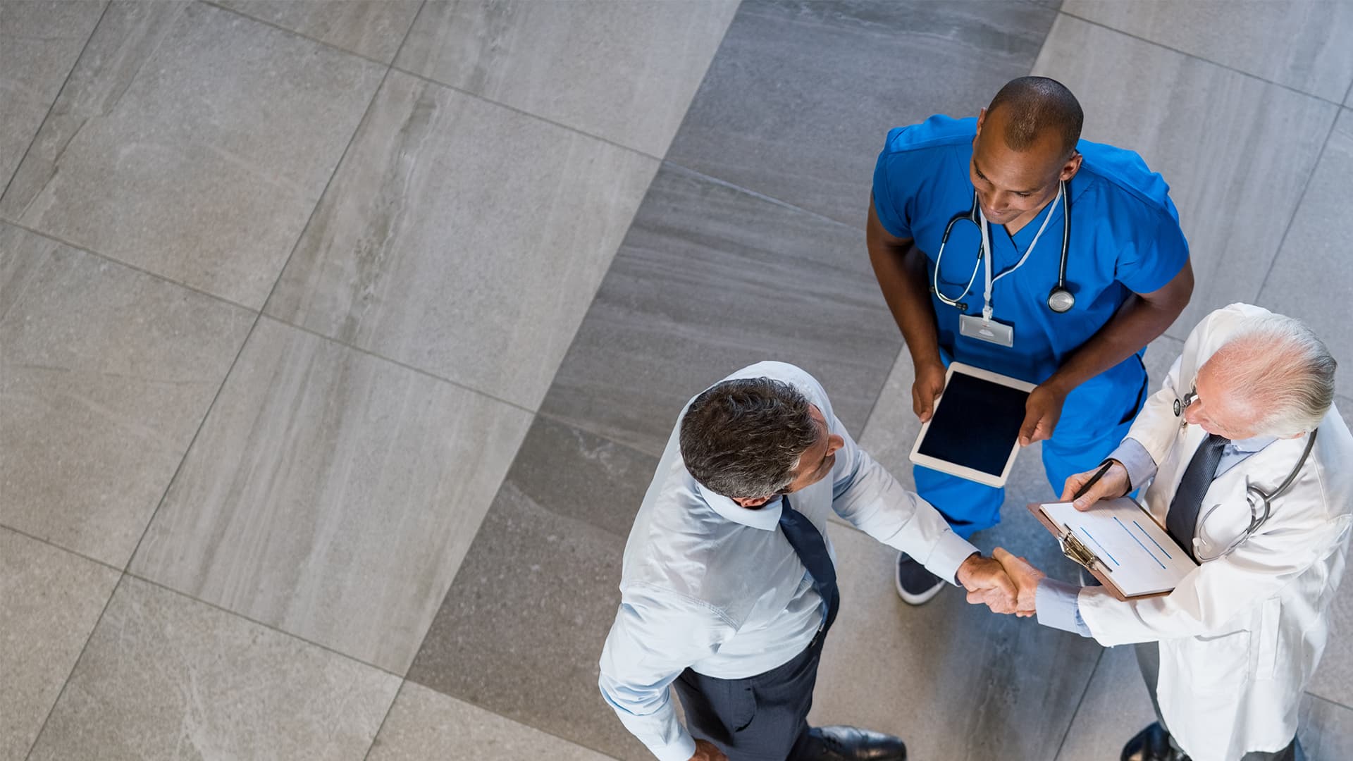 Three men viewed from overhead in a medical setting. Two of them are shaking hands.