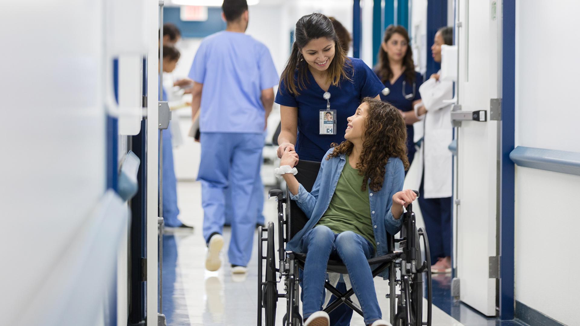 A young girl talks with a nurse, who is pushing the girl in a wheelchair in a busy hospital hallway.
