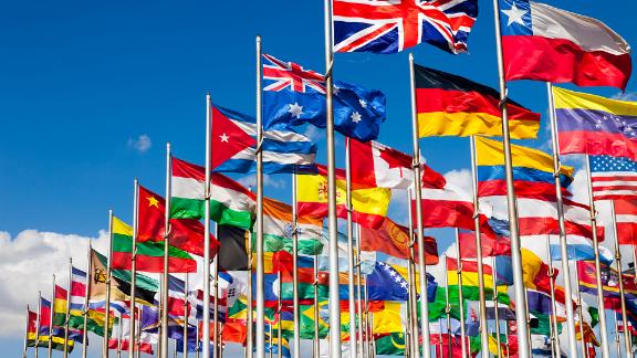 Group of flags of many different nations against blue sky and infront of a convention center.