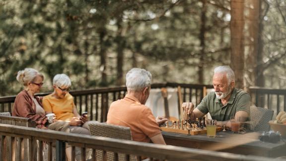 Two men playing a game of chess on a back porch while two women sit beside each other on a bench.