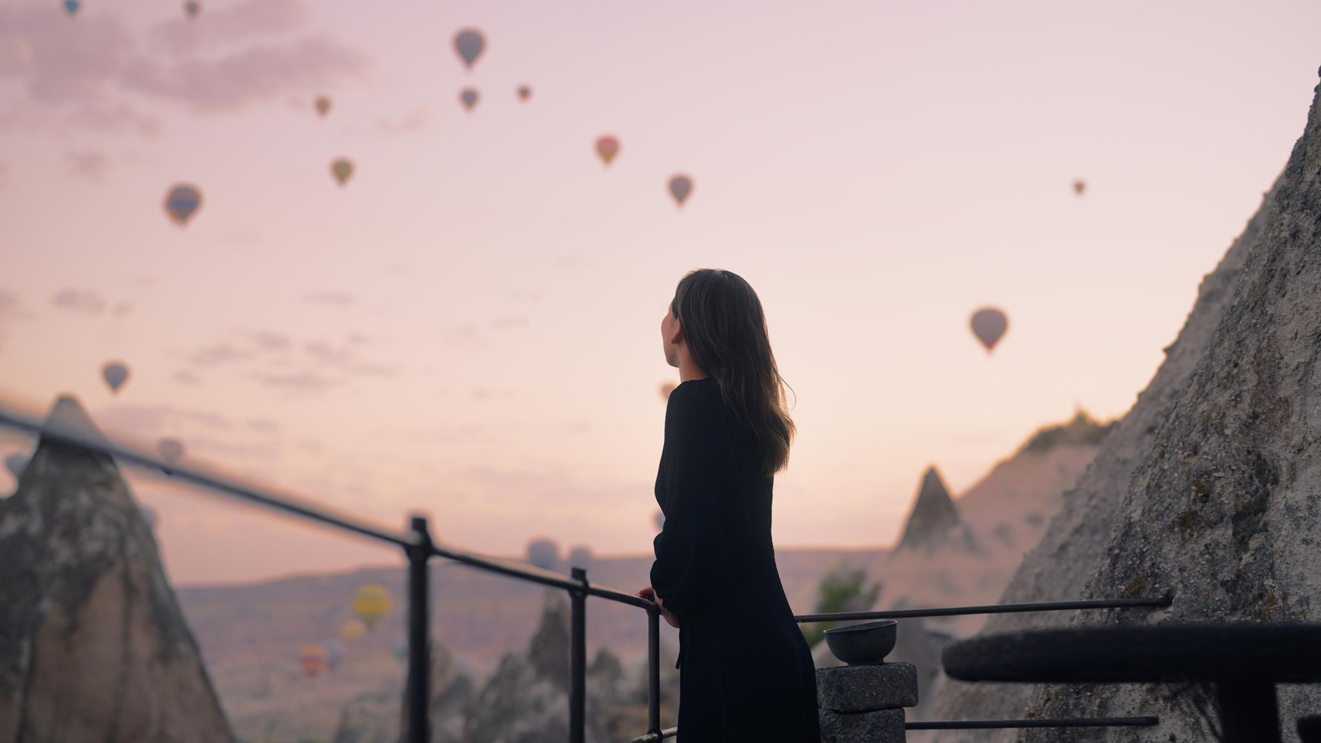 A woman standing on a balcony admiring a hot air balloon festival.